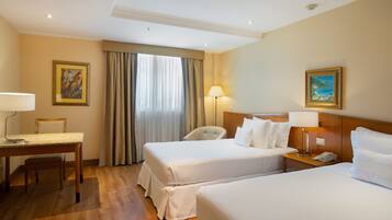 Quarto de hotel com duas camas, uma escrivaninha, uma cadeira, um abajur e um quadro na parede.