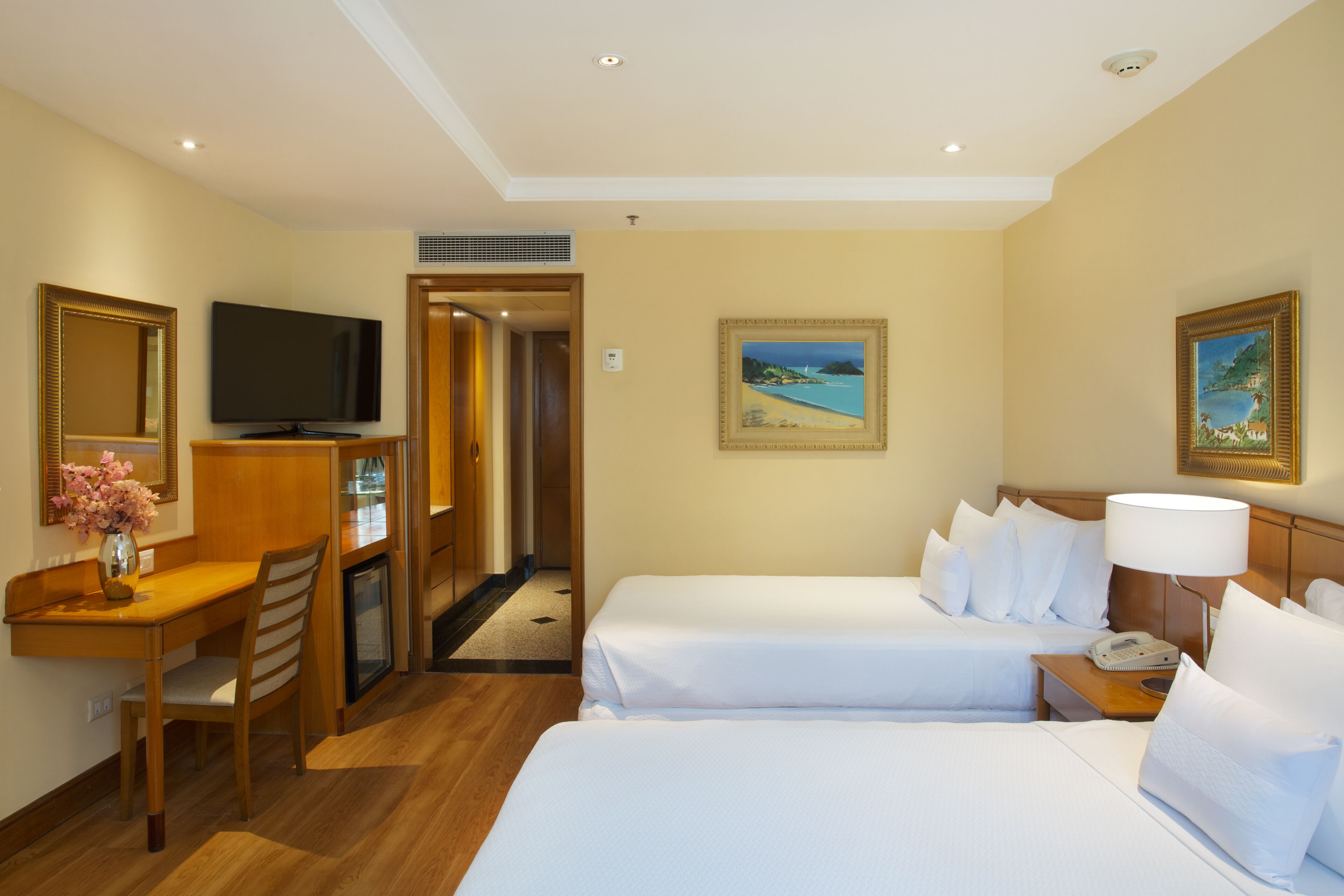 A hotel room with two beds, a desk with a TV, a painting on the wall, and a lamp.