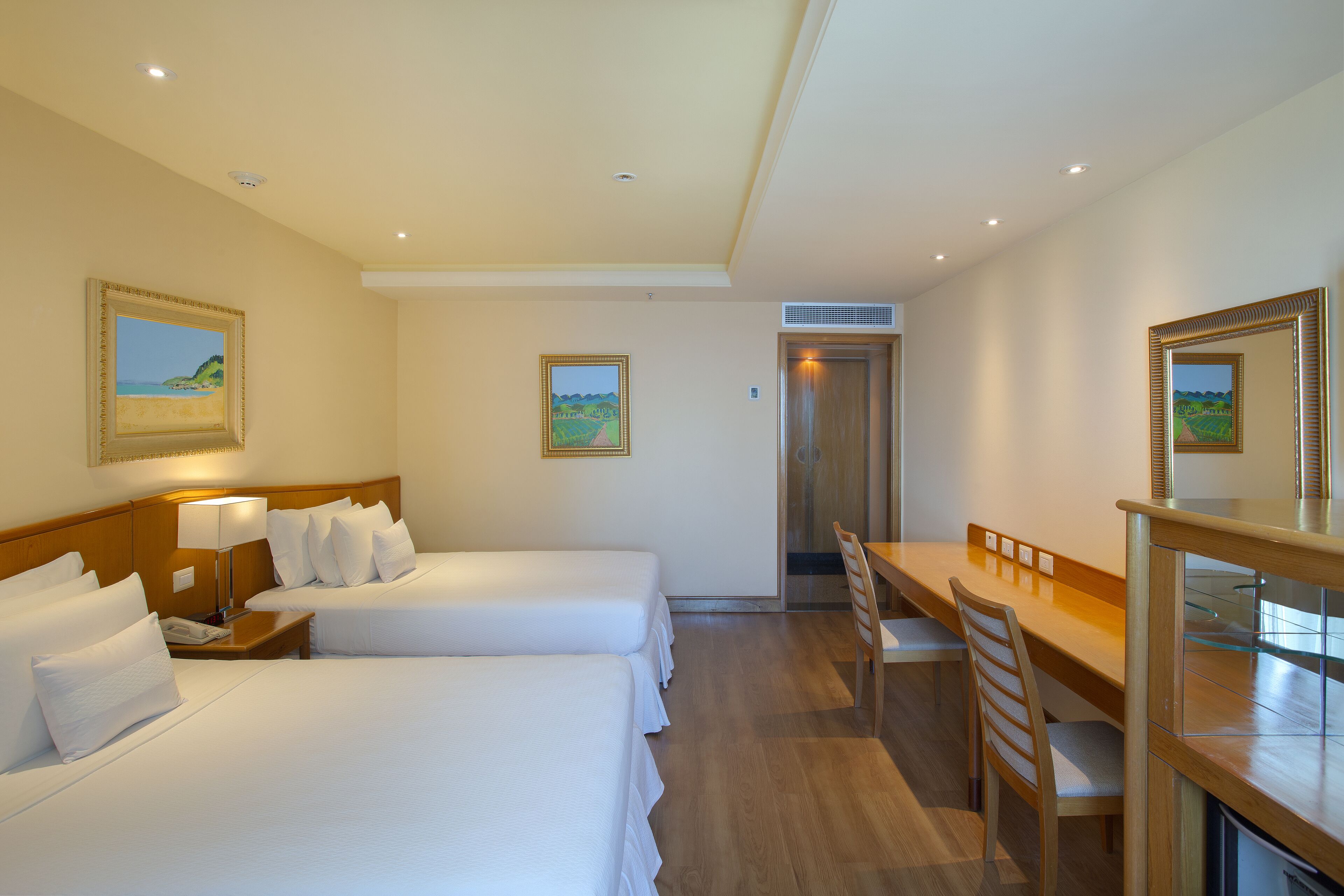 Habitación de hotel con dos camas, un escritorio y una silla. Hay un cuadro en la pared y una puerta que da a otra habitación.