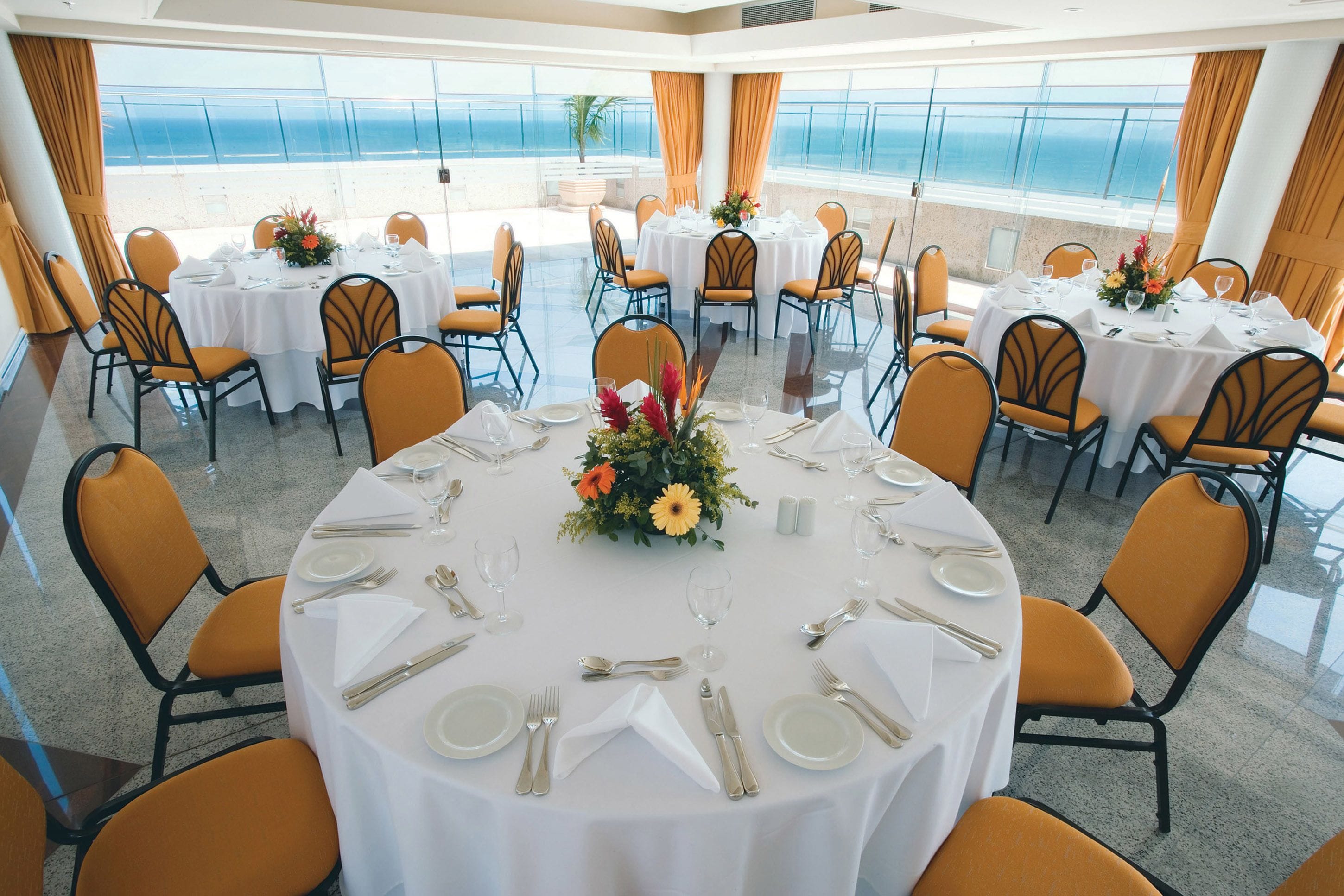 Uma sala de jantar com mesas redondas para seis pessoas, cada uma com toalhas brancas, cadeiras laranja e um arranjo floral central.
