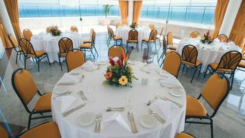 Uma sala de jantar com mesas redondas para seis pessoas, cada uma com toalhas brancas, cadeiras laranja e um arranjo floral central.