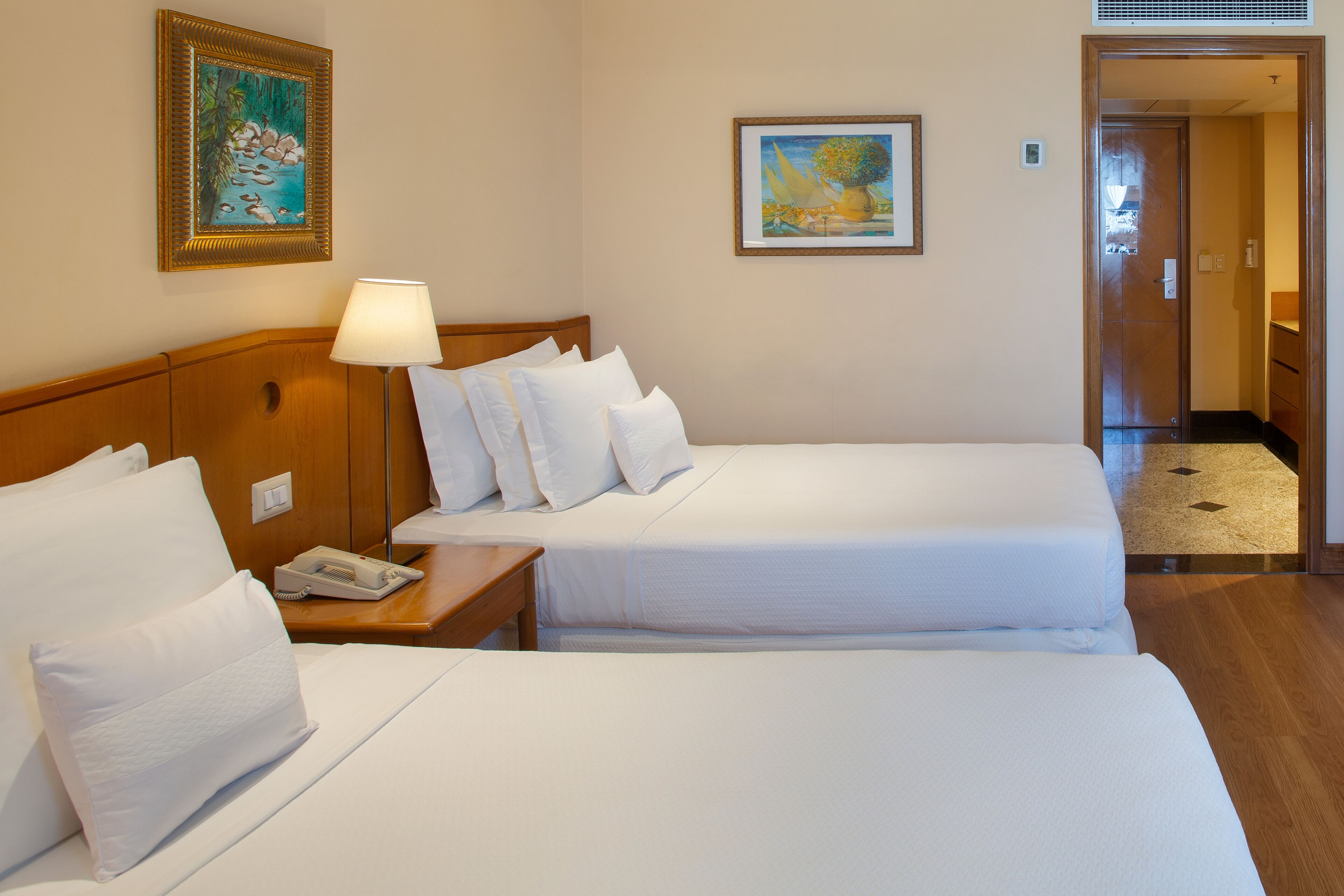 Una habitación de hotel con dos camas, una mesita de noche con lámpara y obras de arte en las paredes.