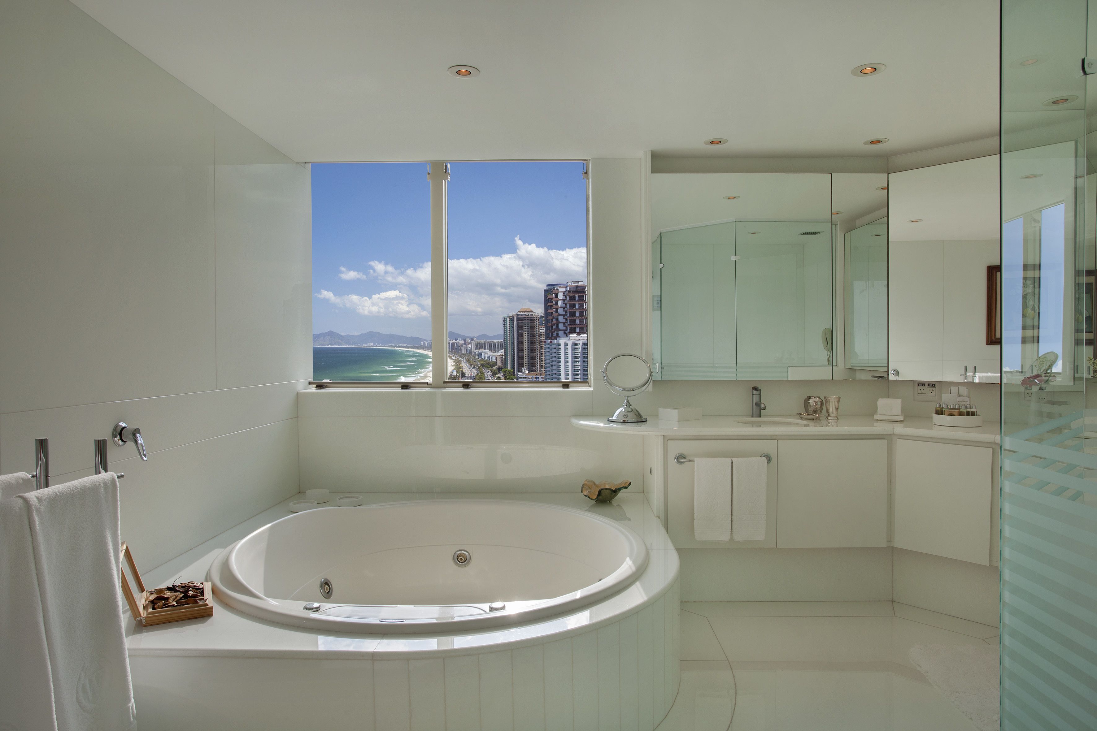 Ein modernes Badezimmer mit einer großen Badewanne, einer Duschkabine aus Glas und Blick auf das Meer und die Stadtlandschaft.