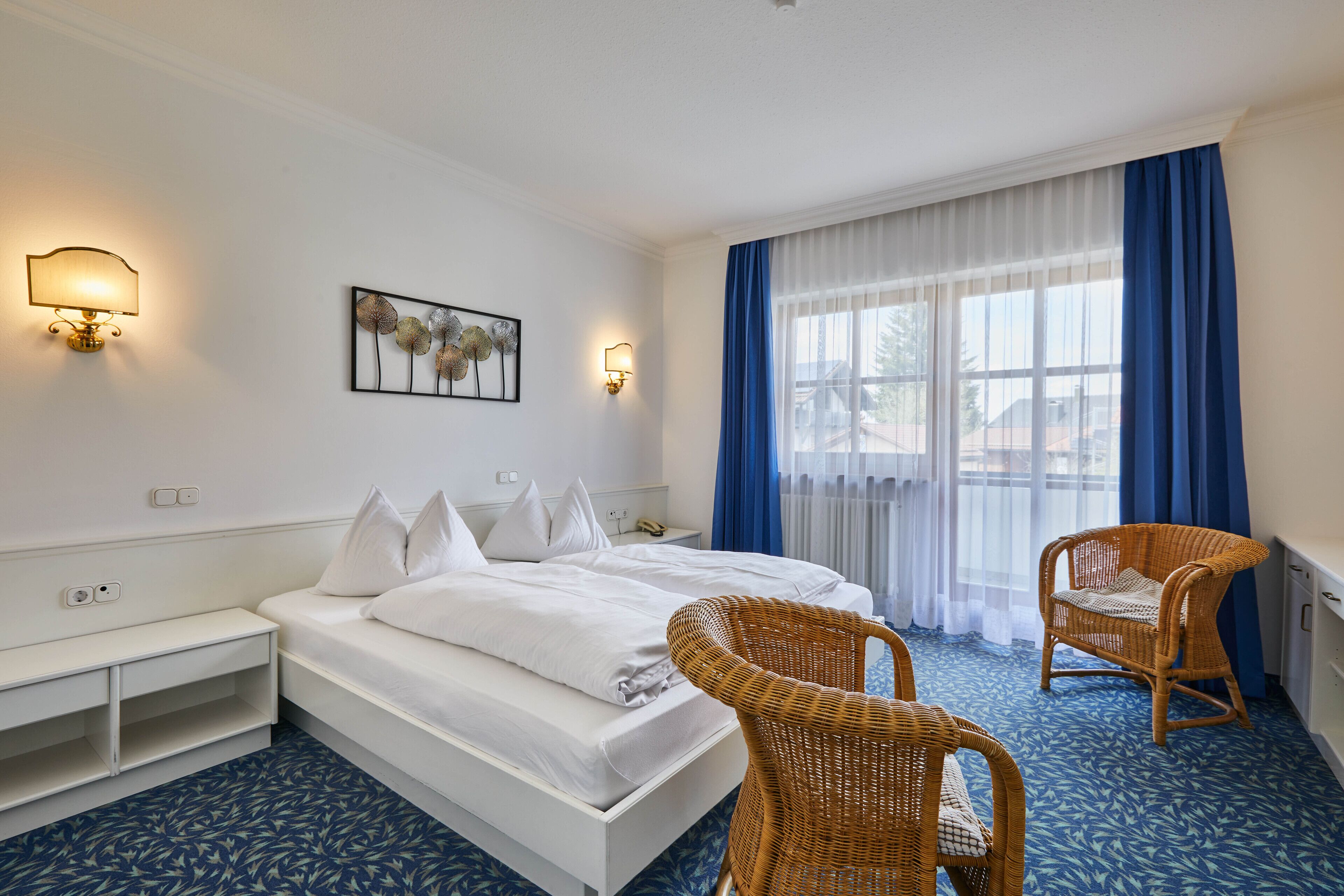 Quarto de hotel com uma cama, duas luminárias fixadas na parede, uma obra de arte emoldurada, uma cadeira de vime e uma janela com cortinas azuis.
