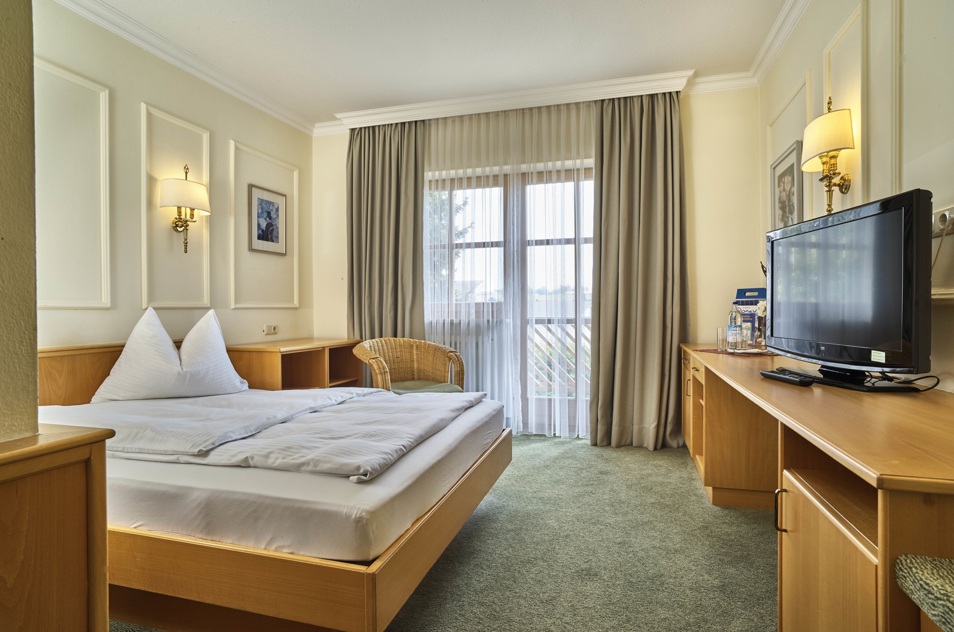 Habitación de hotel con una cama grande, un escritorio con televisión, una silla y una ventana con cortinas.