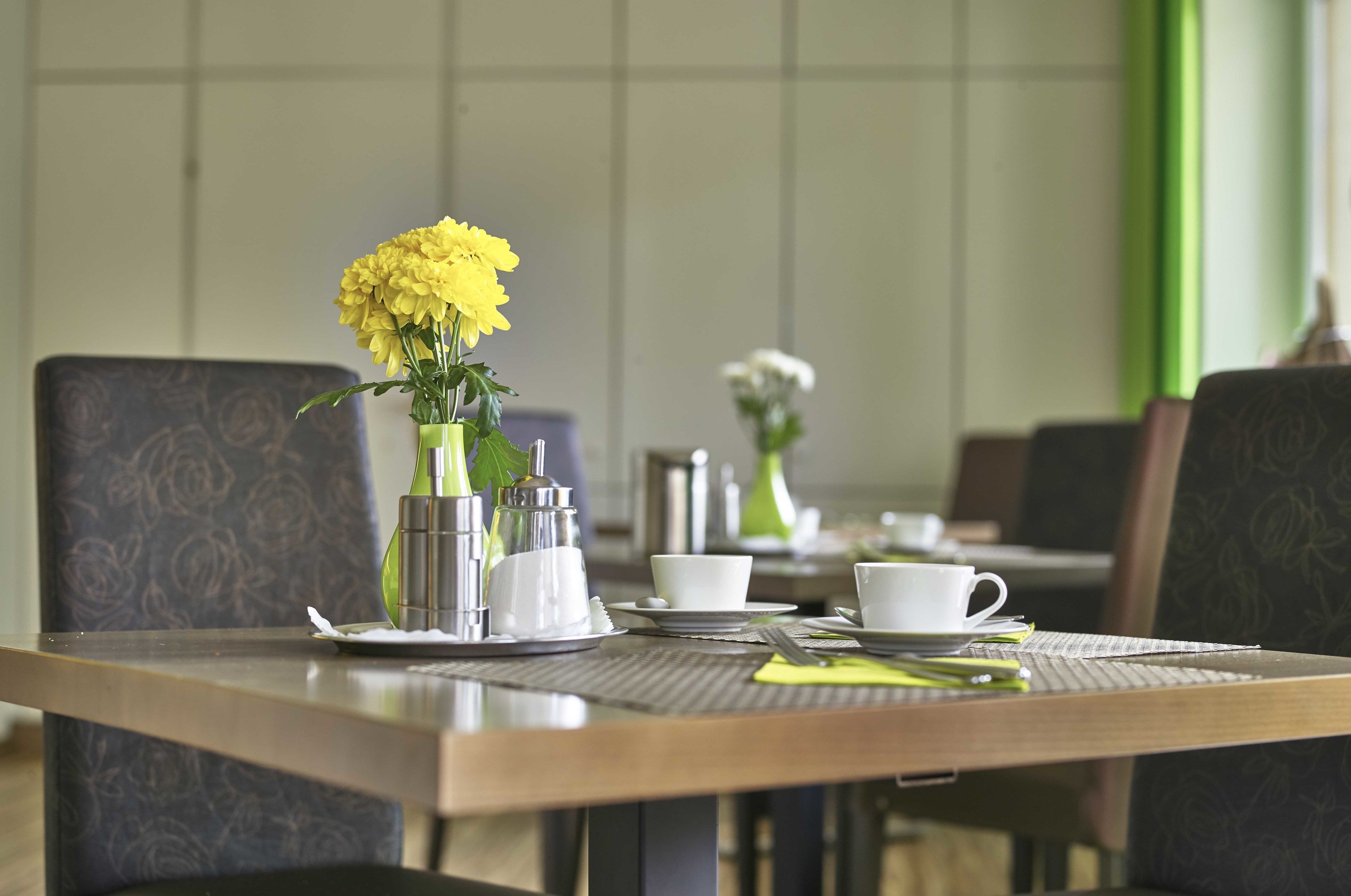 A dining table set with a vase of yellow flowers, white cups, and a green vase.