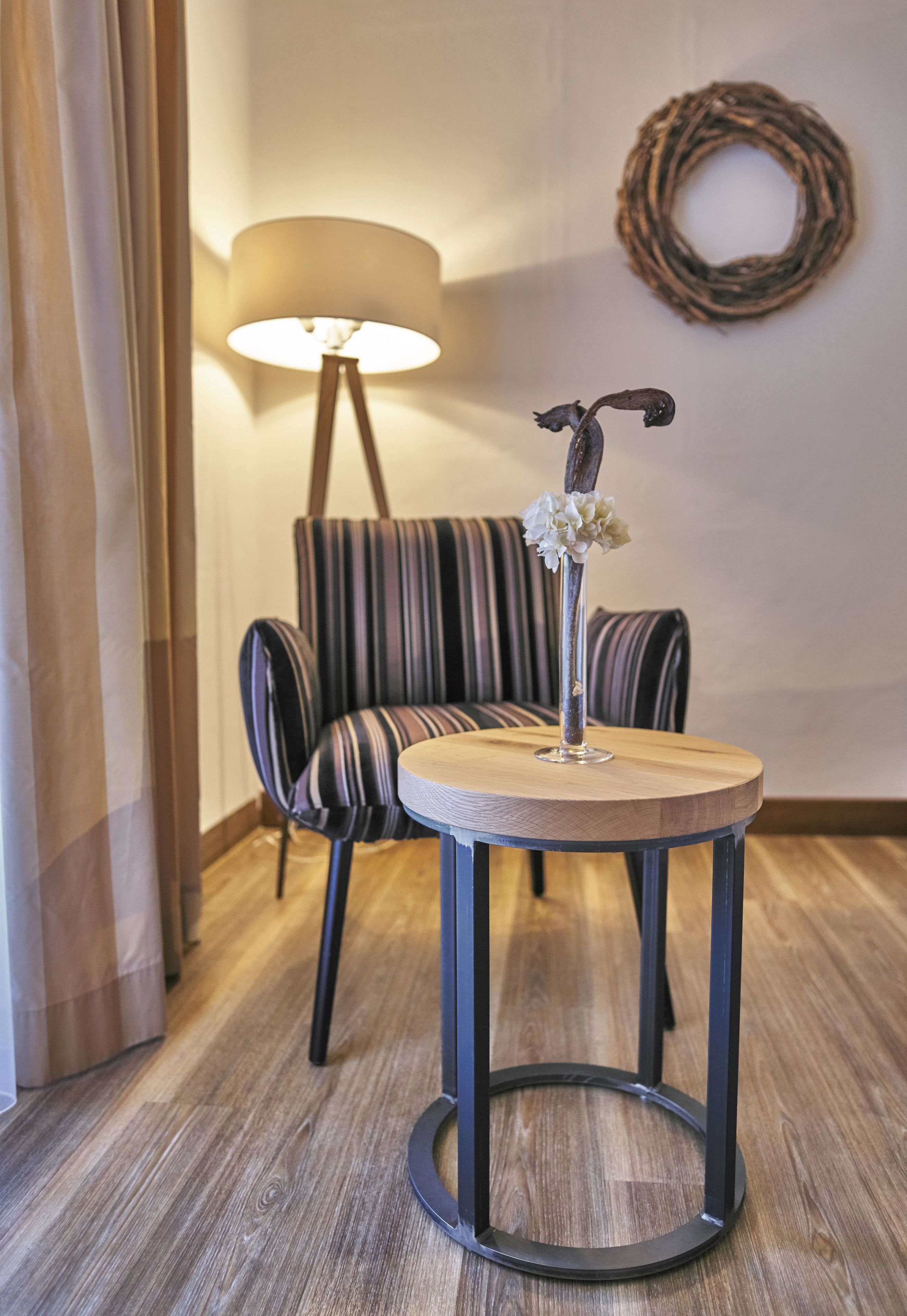 Ein runder Holztisch mit Metallfuß, ein gestreifter Sessel, eine Stehlampe und eine Wandkrans.