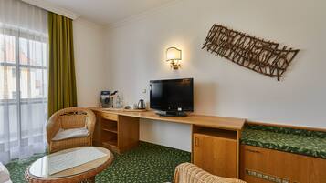 Habitación de hotel con televisión, muebles de mimbre, alfombra estampada en verde y una obra de arte colgada en la pared.
