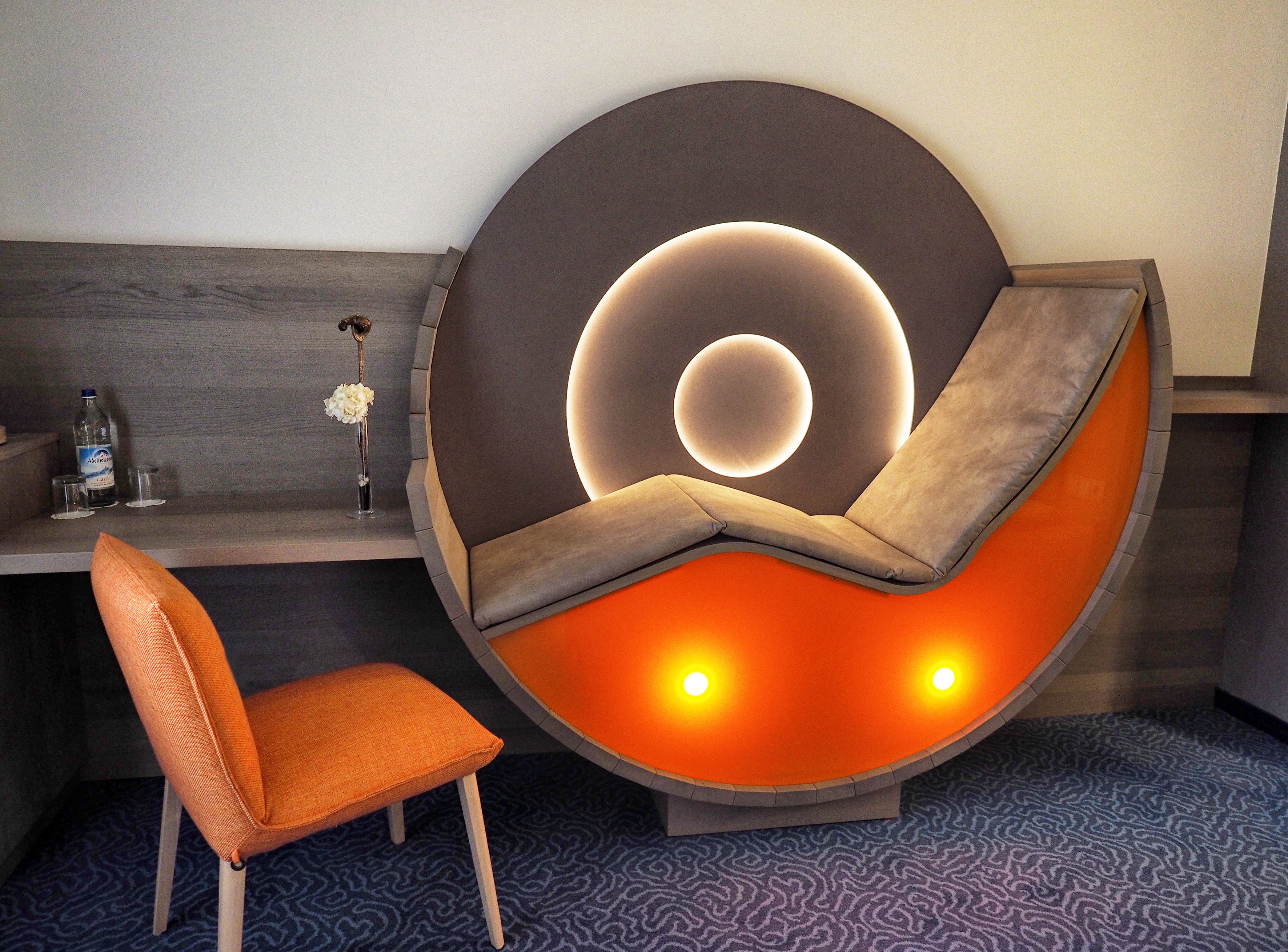 Ein modernes Zimmer mit einer einzigartigen, runden Sitzecke, einem kleinen Tisch mit einer Vase und Flaschen sowie einem gemusterten Teppich.