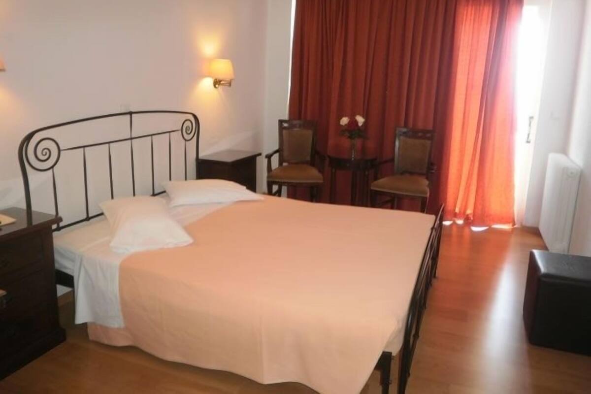 A neatly made bed with a headboard, a bedside table, a chair, a small table with a vase of flowers, and a red curtain.