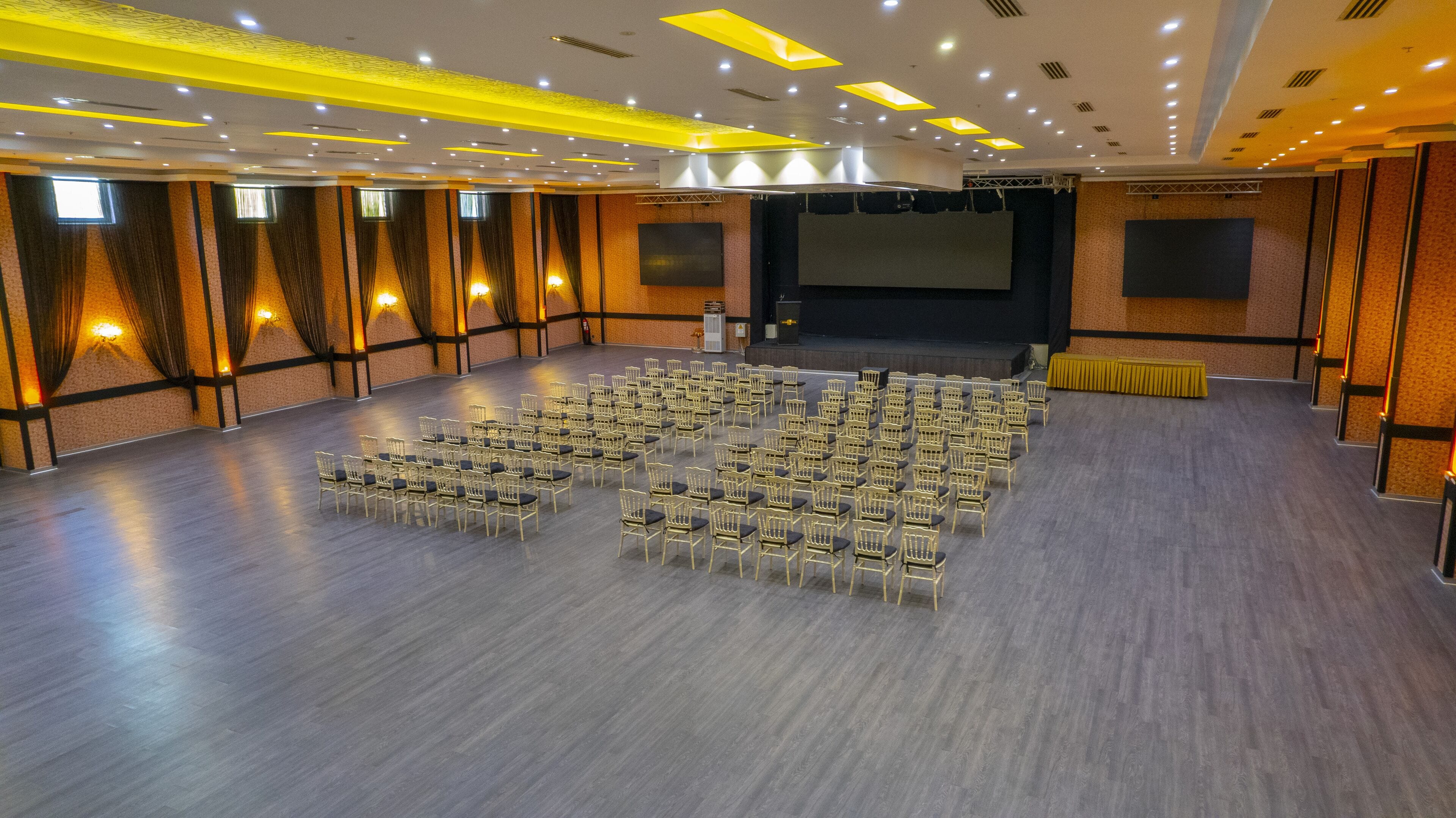 Une grande salle avec des rangées de chaises disposées pour un événement, une scène avec un écran et un plafond doté d’un éclairage encastré.