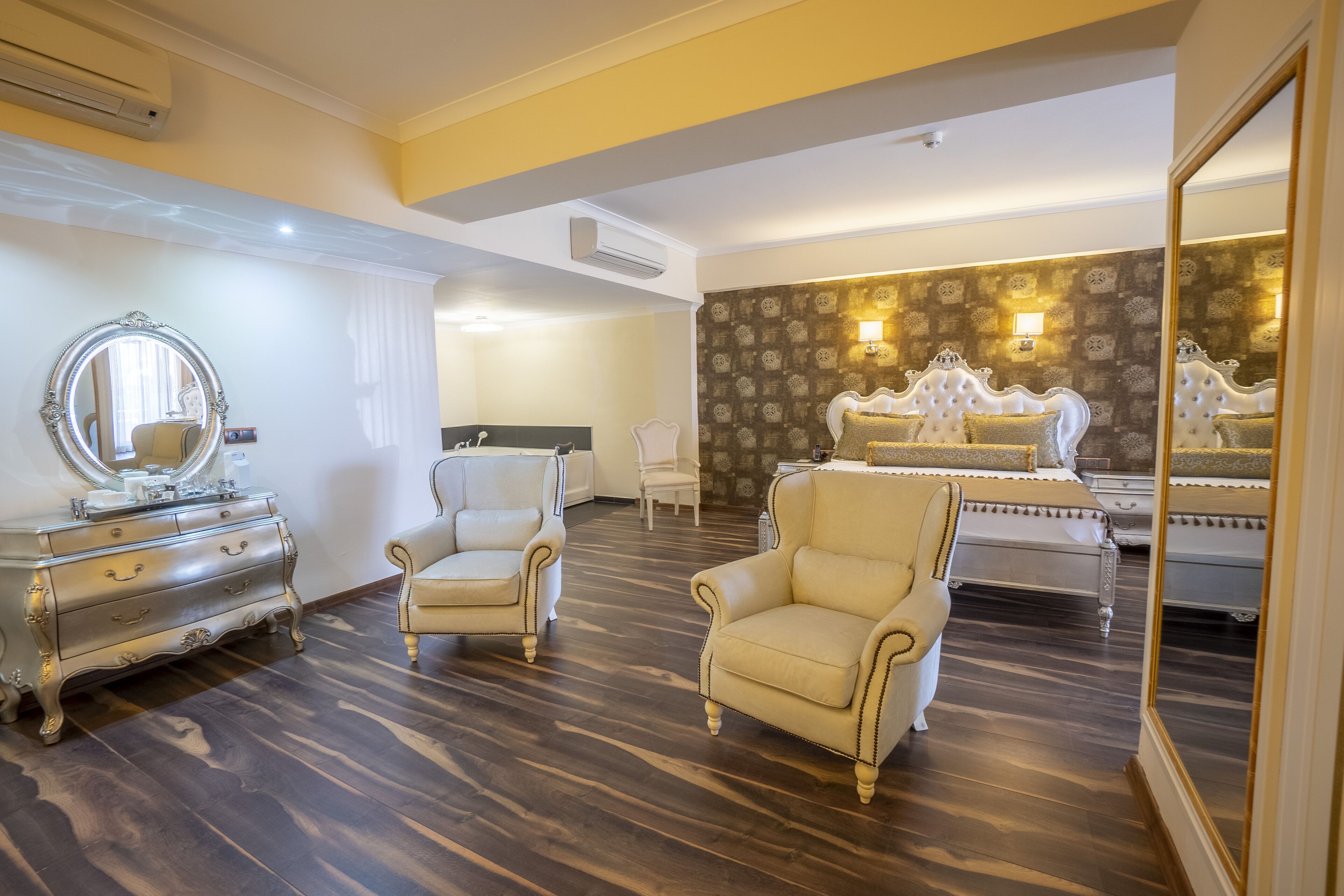 A hotel room with a large bed, a vanity with a mirror, two armchairs, and a dresser.