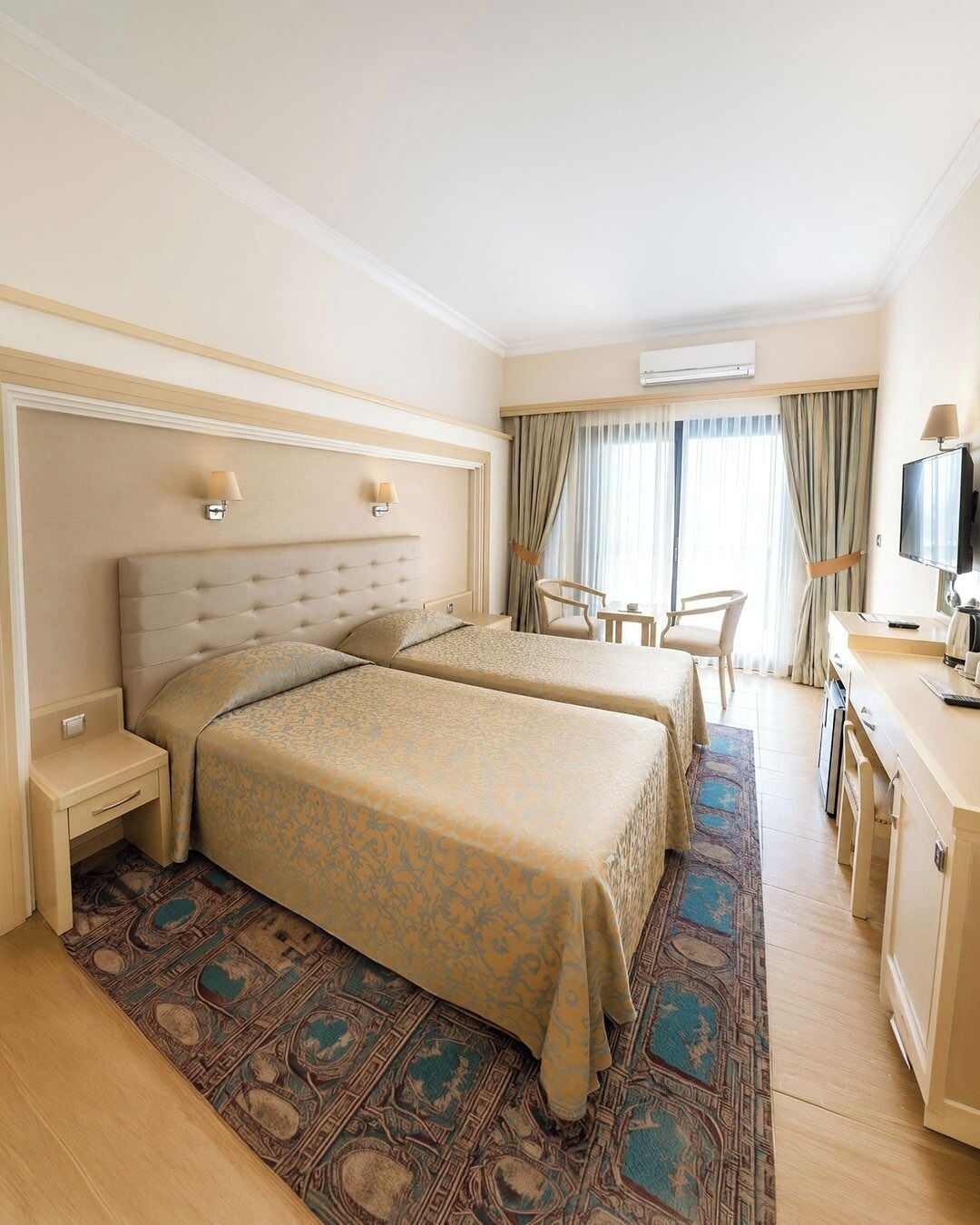 A hotel room with two beds, a desk, a chair, a television, and an air conditioning unit.