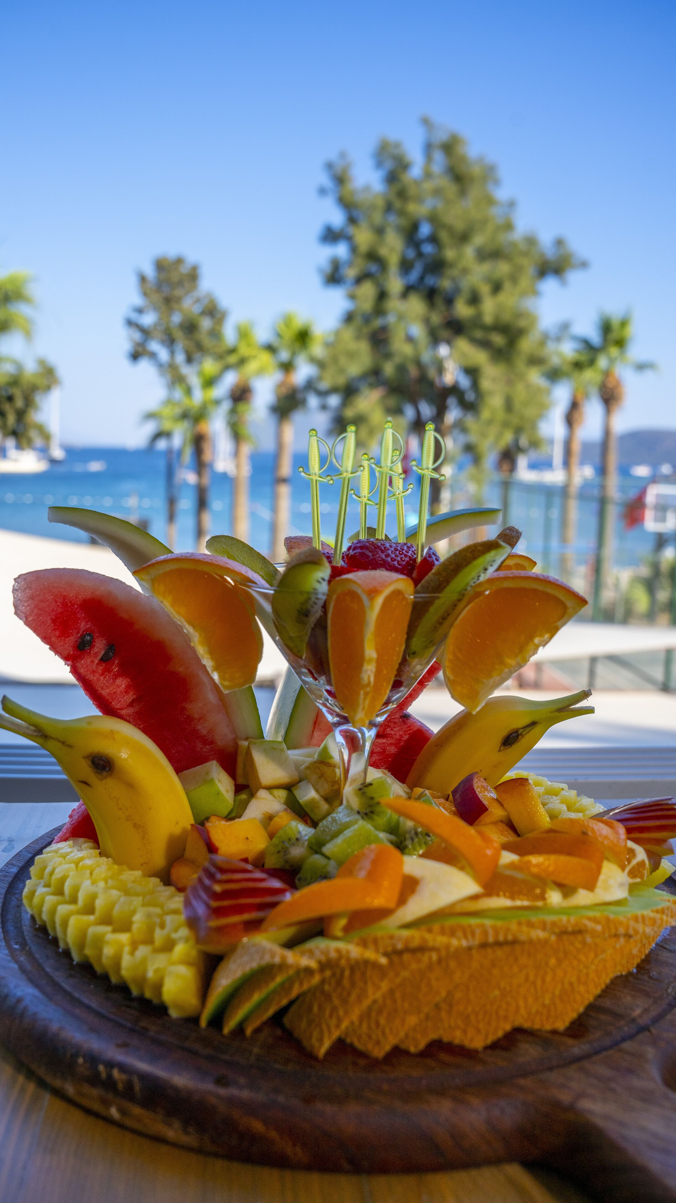 A fruit platter with a variety of fruits and a slice of bread on a wooden board, placed on a table with a view of the ocean and palm trees in the background.
