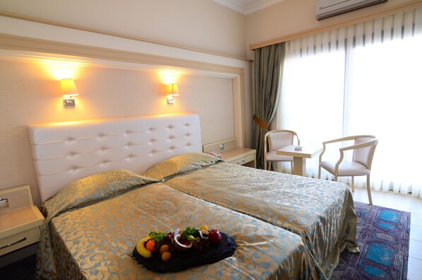 A hotel room with two beds, a small desk, a chair, and a fruit basket on the bed. - Azka Hotel (Bodrum)