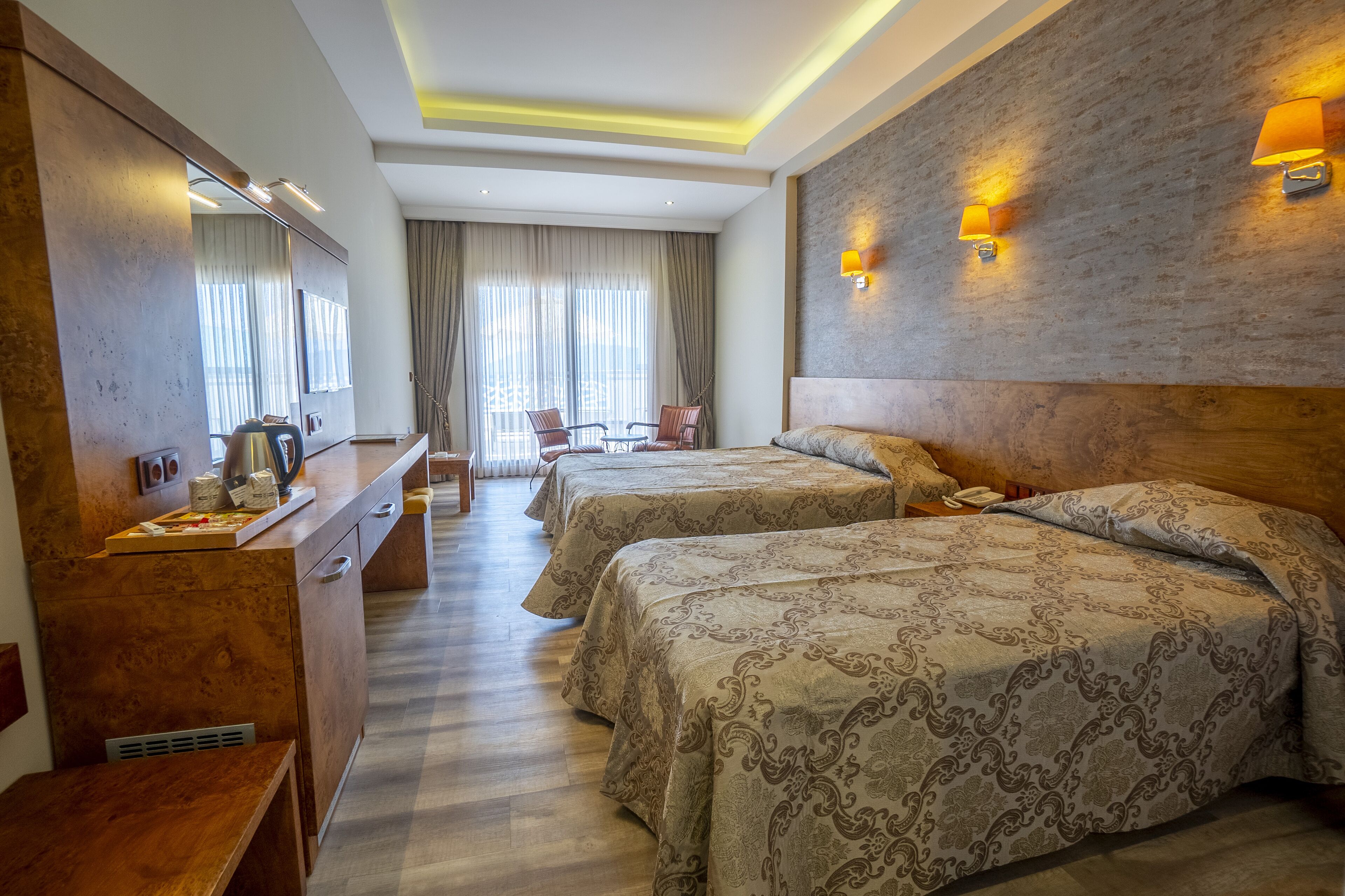 A hotel room with two beds, a wooden desk, a mirror, and a view of the outdoors.