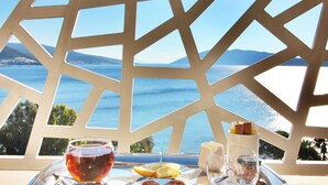 Una mesa con bandeja dispuesta para una comida, que incluye una copa de vino tinto, una copa de agua y algunos pasteles, con una vista panorámica del mar y las montañas al fondo.