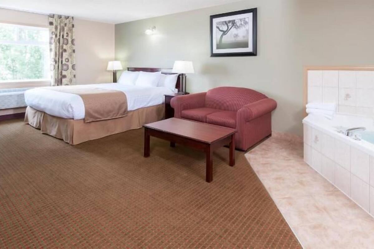A hotel room with two beds, a red sofa, a small table, a bathtub, and a window with curtains.
