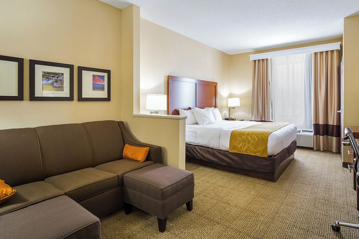 A hotel room with a large bed, a sofa, and a small table. There are framed pictures on the wall and a window with curtains.
