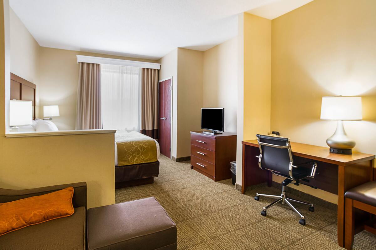 A hotel room with a bed, a desk with a chair, a television, a dresser, and a window with curtains.
