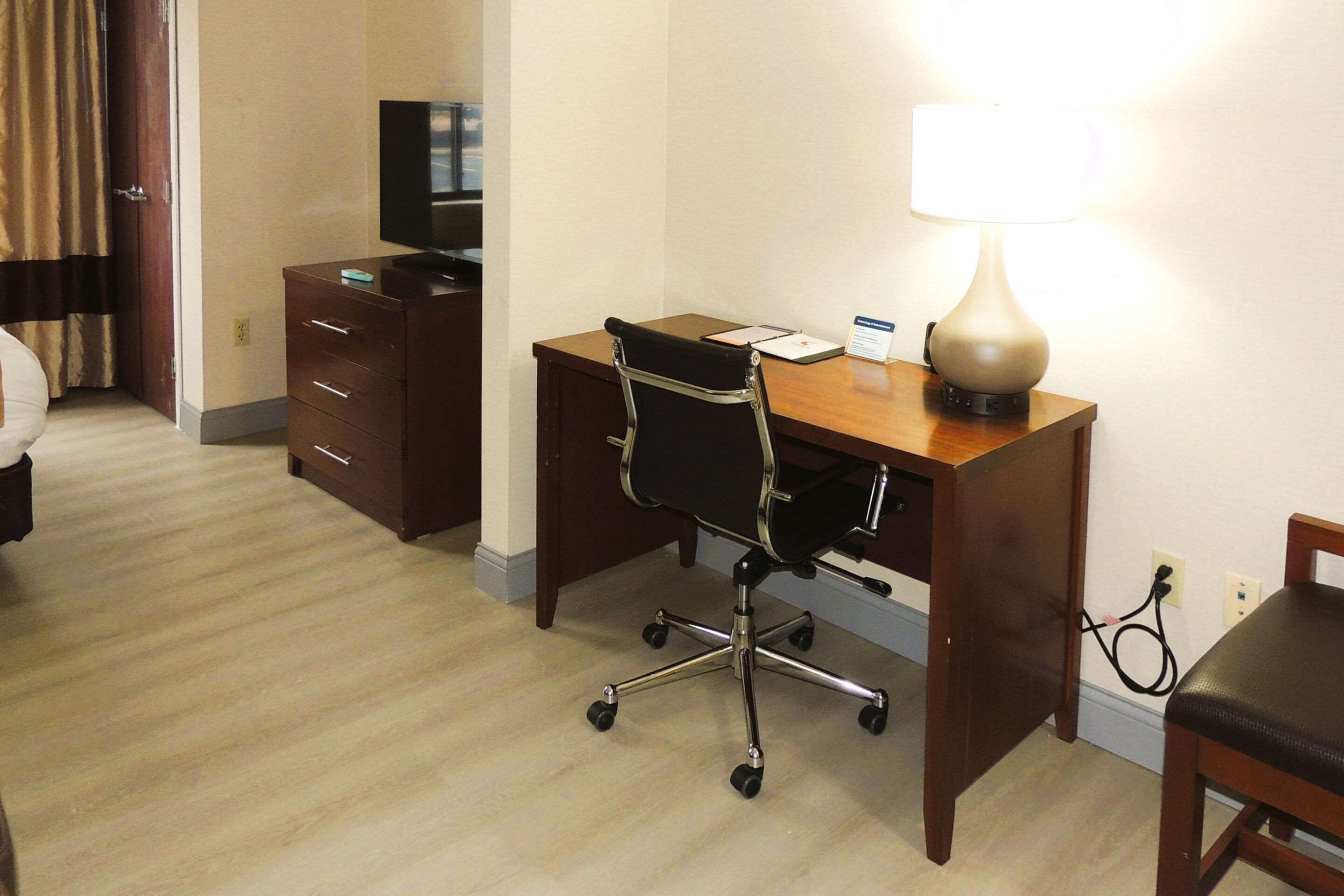 Quarto de hotel com escrivaninha de madeira, cadeira de escritório preta, luminária, televisão e cama.