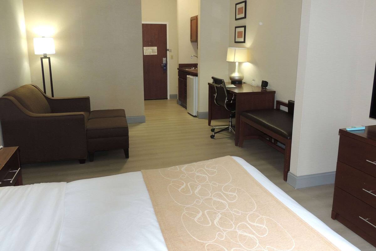 A hotel room with a bed, a desk, a chair, a lamp, a dresser, and a kitchenette in the background.