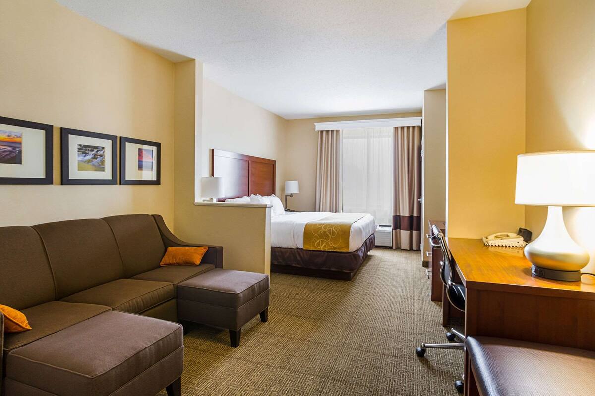 A hotel room with a bed, a sofa, an ottoman, a desk with a lamp, and framed pictures on the wall.