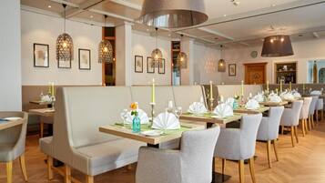 A restaurant with multiple dining tables, grey upholstered booths, and pendant lighting.