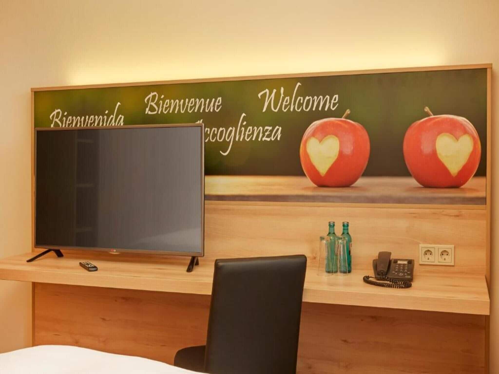 A hotel reception desk with a flat-screen TV, a wooden desk, a black chair, and a wall with a welcome sign and two apples with heart designs.