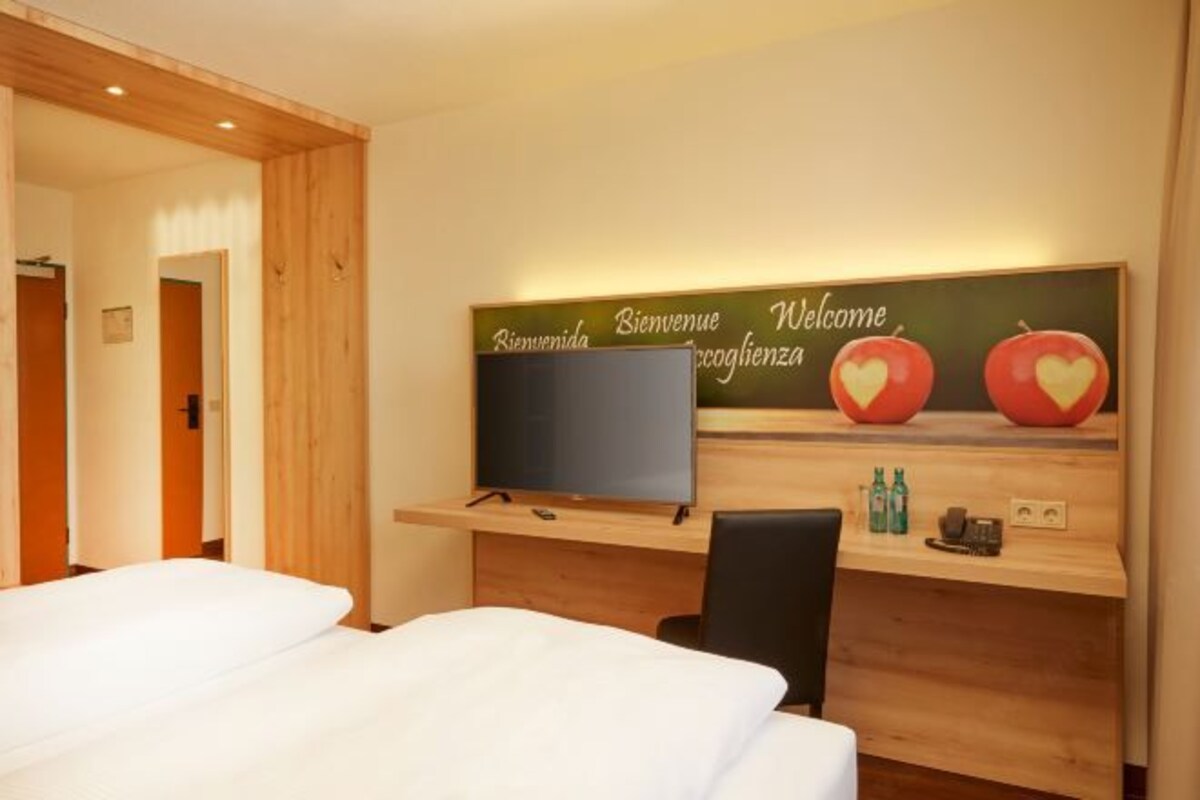 A hotel room with a bed, a desk with a flat-screen TV, and a reception desk with a welcome sign.