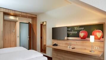 A hotel room with two beds, a wooden wall, a welcome sign, and a small desk with a phone and glasses.