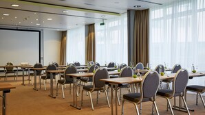 Ein Konferenzraum mit mehreren Tischen und Stühlen, großen Fenstern mit Vorhängen und einer Projektionsleinwand.