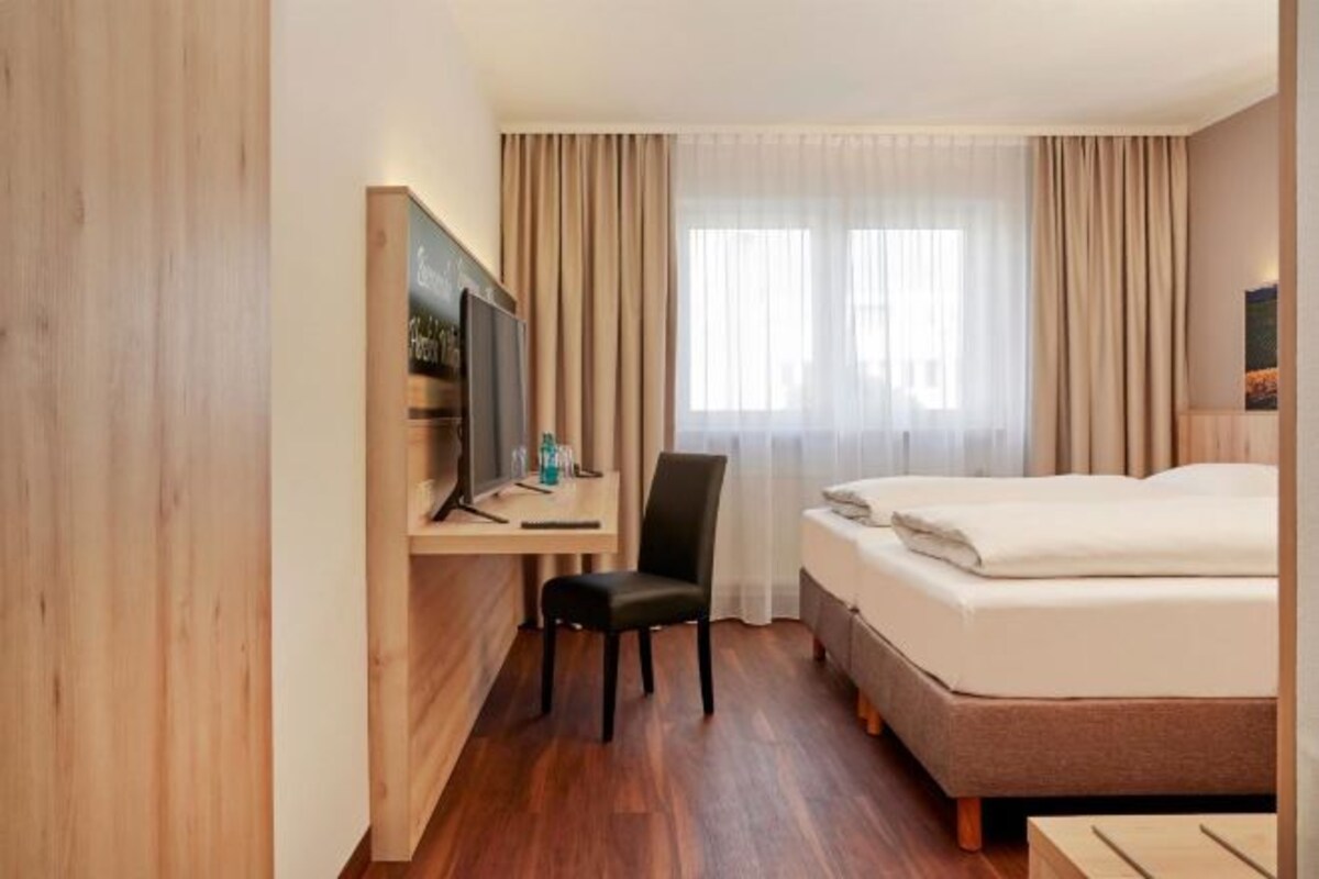 A hotel room with a bed, a desk with a TV, a chair, and a window with curtains.