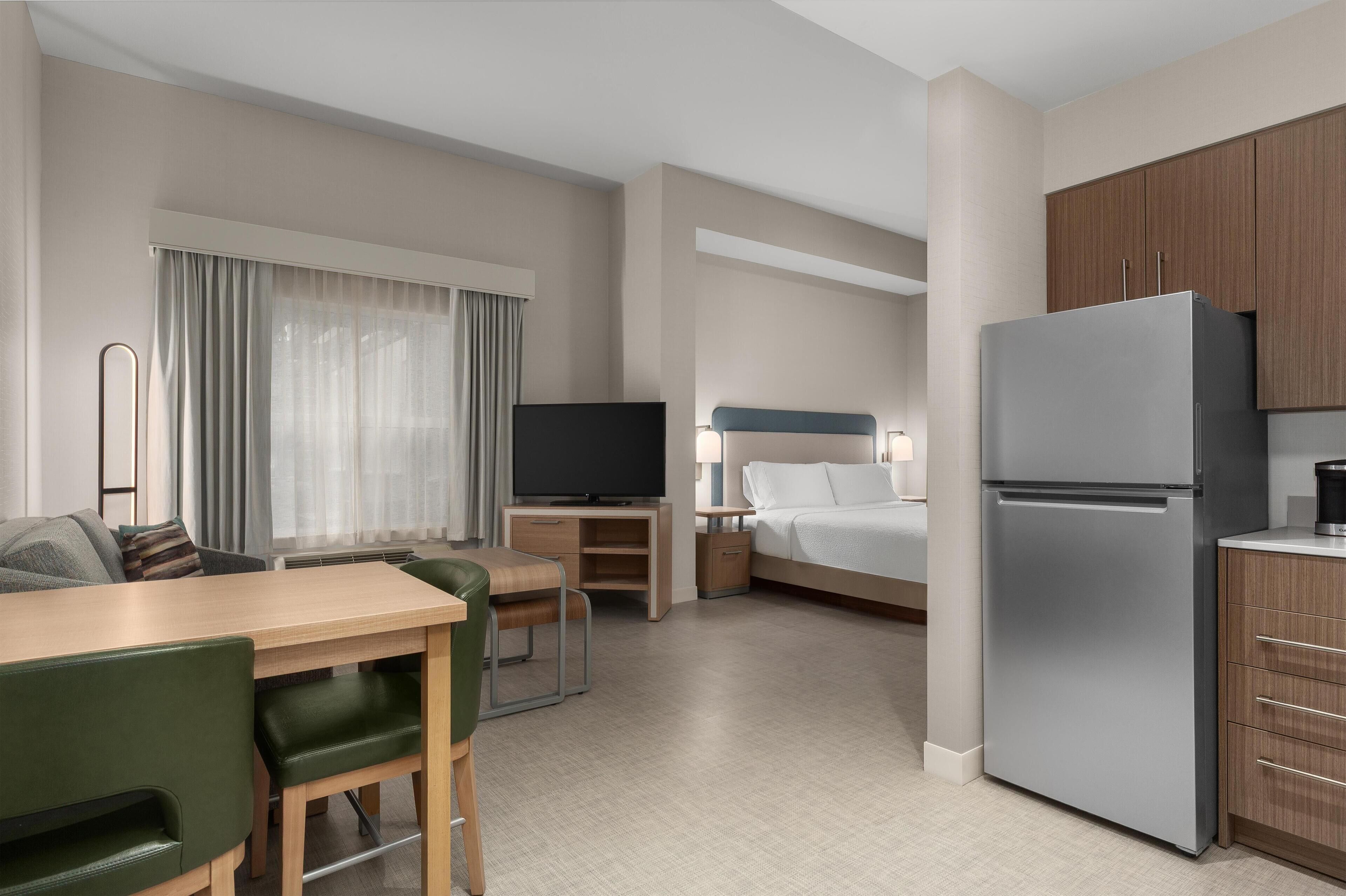 Habitación de hotel con comedor, cama, televisor, refrigerador y kitchenette.
