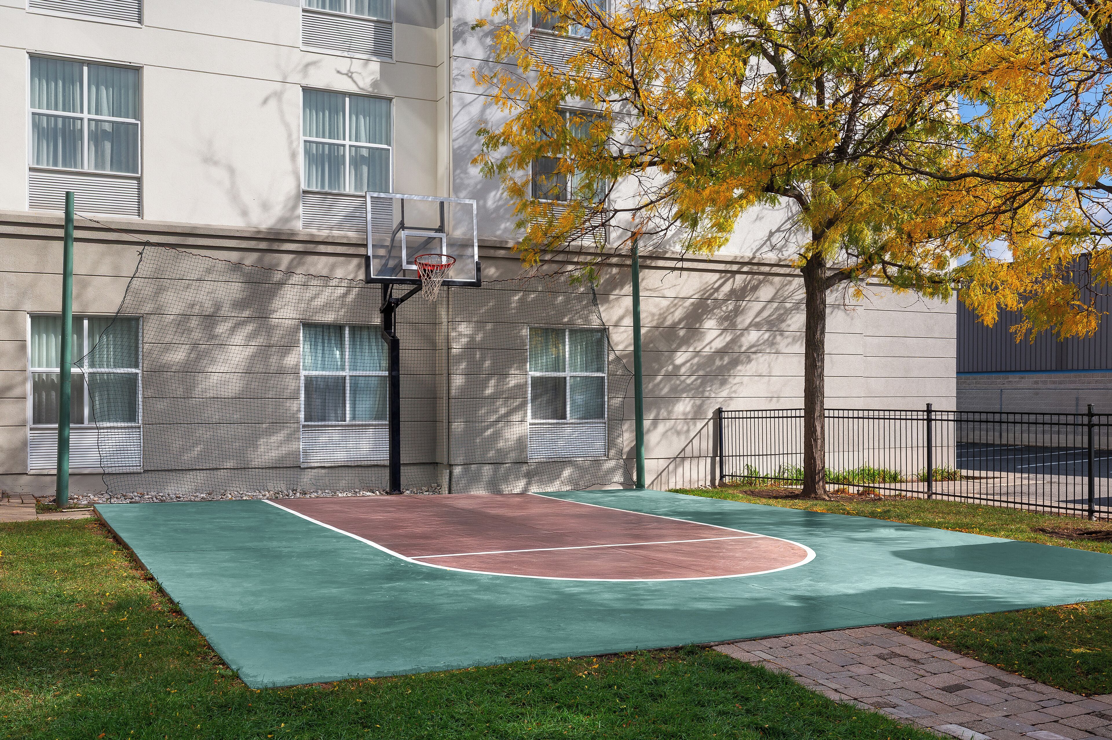 Una cancha de baloncesto al aire libre junto a un edificio con ventanas y un árbol con hojas amarillas.