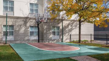 Una cancha de baloncesto al aire libre junto a un edificio con ventanas y un árbol con hojas amarillas.