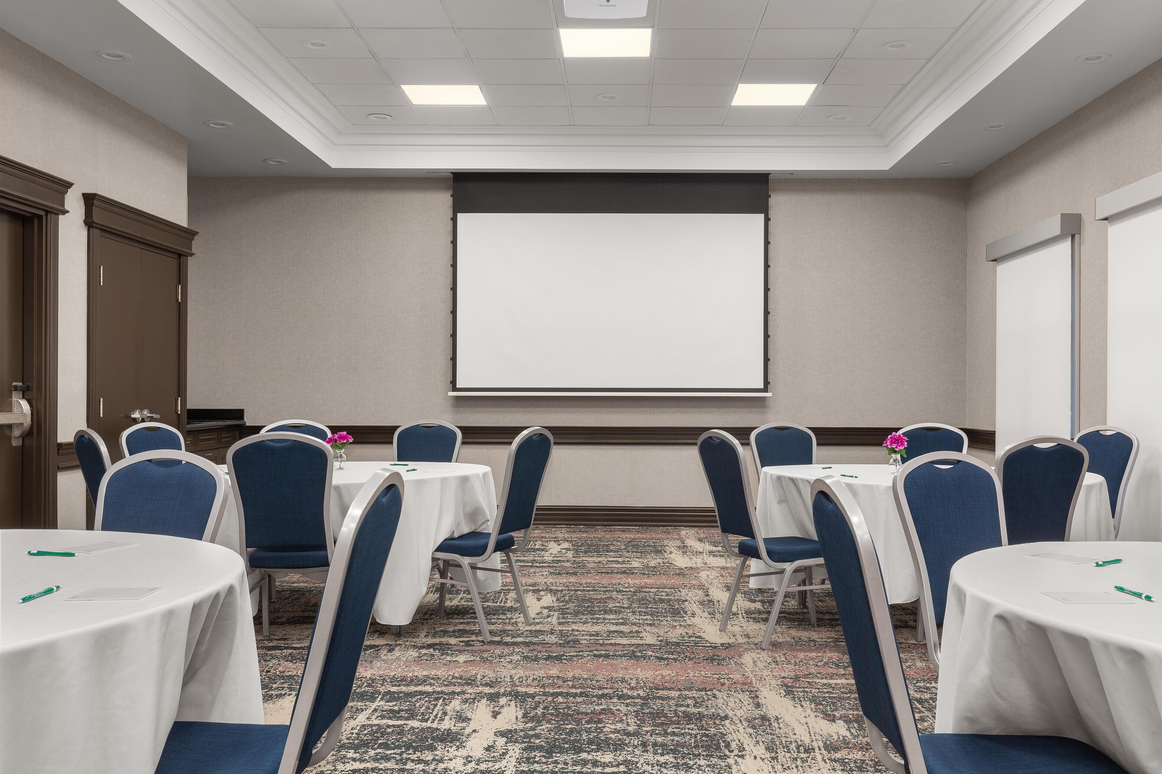 Sala de conferências com mesas brancas, cadeiras azuis, tela de projetor e piso carpeteado.