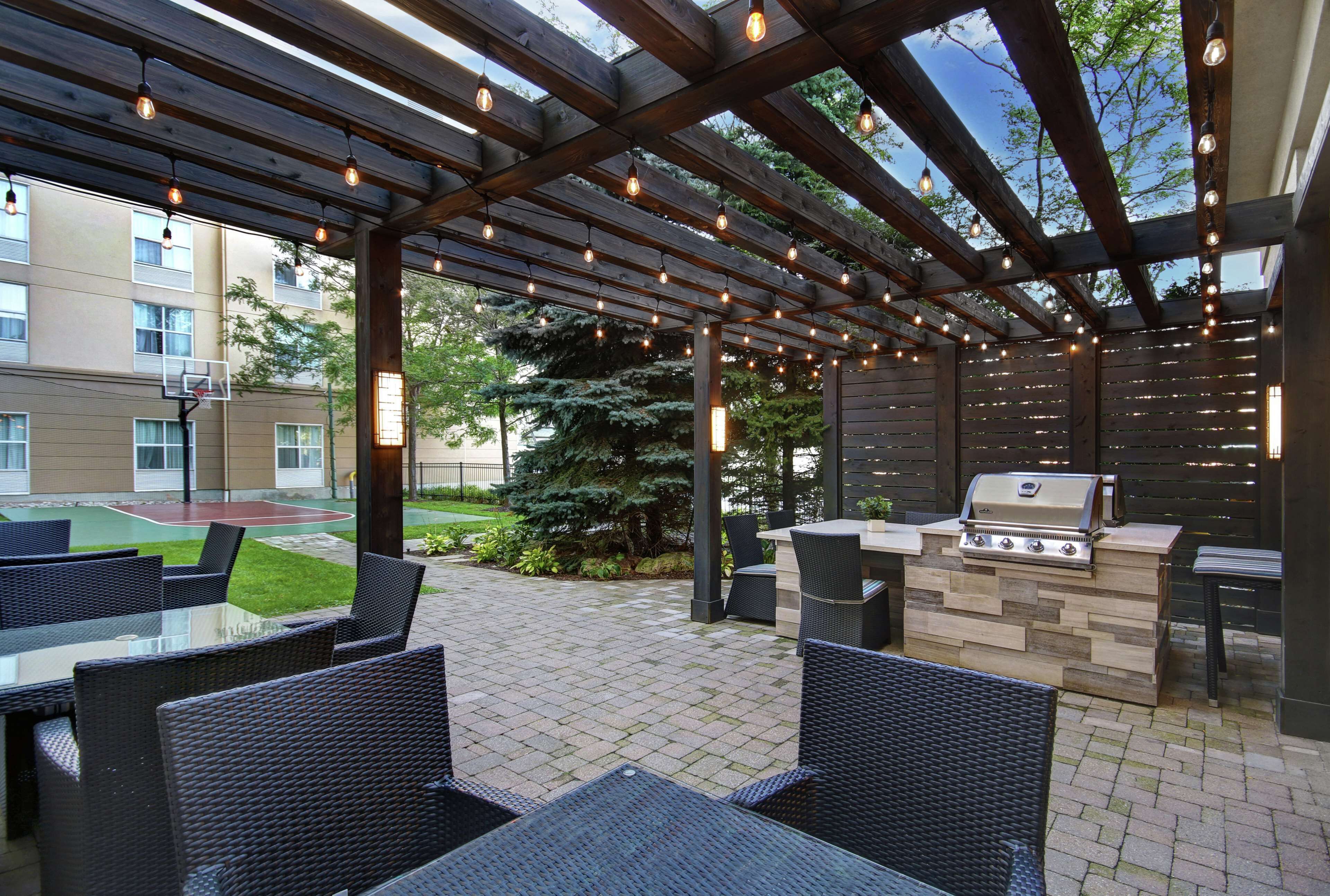 Un patio exterior con una pérgola de madera, luces colgantes, una parrilla y mobiliario de mimbre.