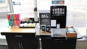 Een koffiehoek met een koffieapparaat, een presentatie van koffiekopjes en een stapel koffieboxen.