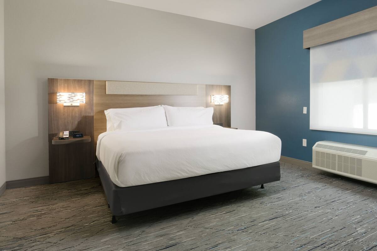 A modern hotel room with a large bed, bedside lamps, a nightstand, and a window with blinds.