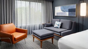 A modern hotel room with a grey sofa, an orange armchair, a small wooden table, and a large window with sheer curtains. - Courtyard by Marriott Kingston (Kingston)