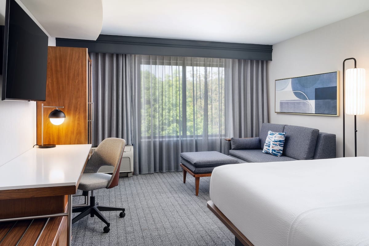 A hotel room with a bed, a desk, a chair, a sofa, a TV, and a window with curtains.