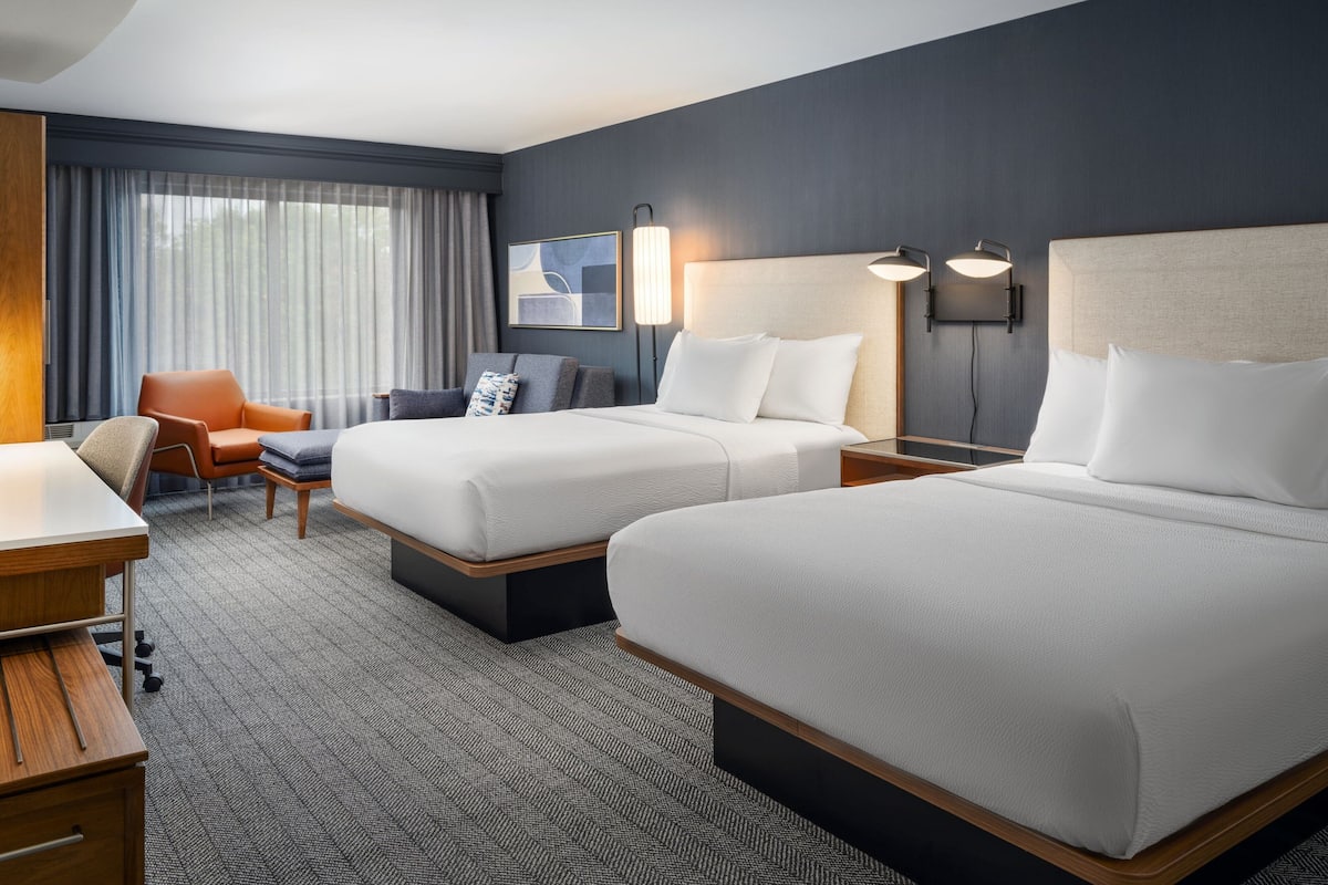 A hotel room with two beds, a desk, a chair, and a large window with curtains.