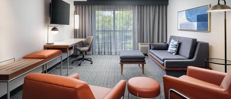 A hotel room with a desk, two orange chairs, a grey sofa, a TV, and a large window with curtains.