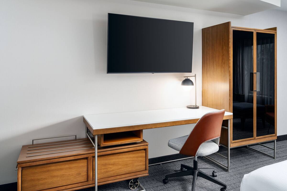 A hotel room with a wooden desk, a chair, a television mounted on the wall, and a wardrobe with a mirror.