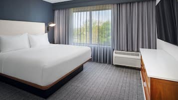 A hotel room with a large bed, a desk, a window with curtains, and an air conditioning unit.
