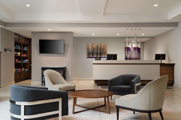 A hotel lobby with a reception desk, two armchairs, a coffee table, and a TV mounted on the wall.