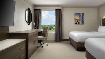 Habitación de hotel con escritorio, dos camas, una silla, un ventanal con cortinas y un cuadro en la pared.
