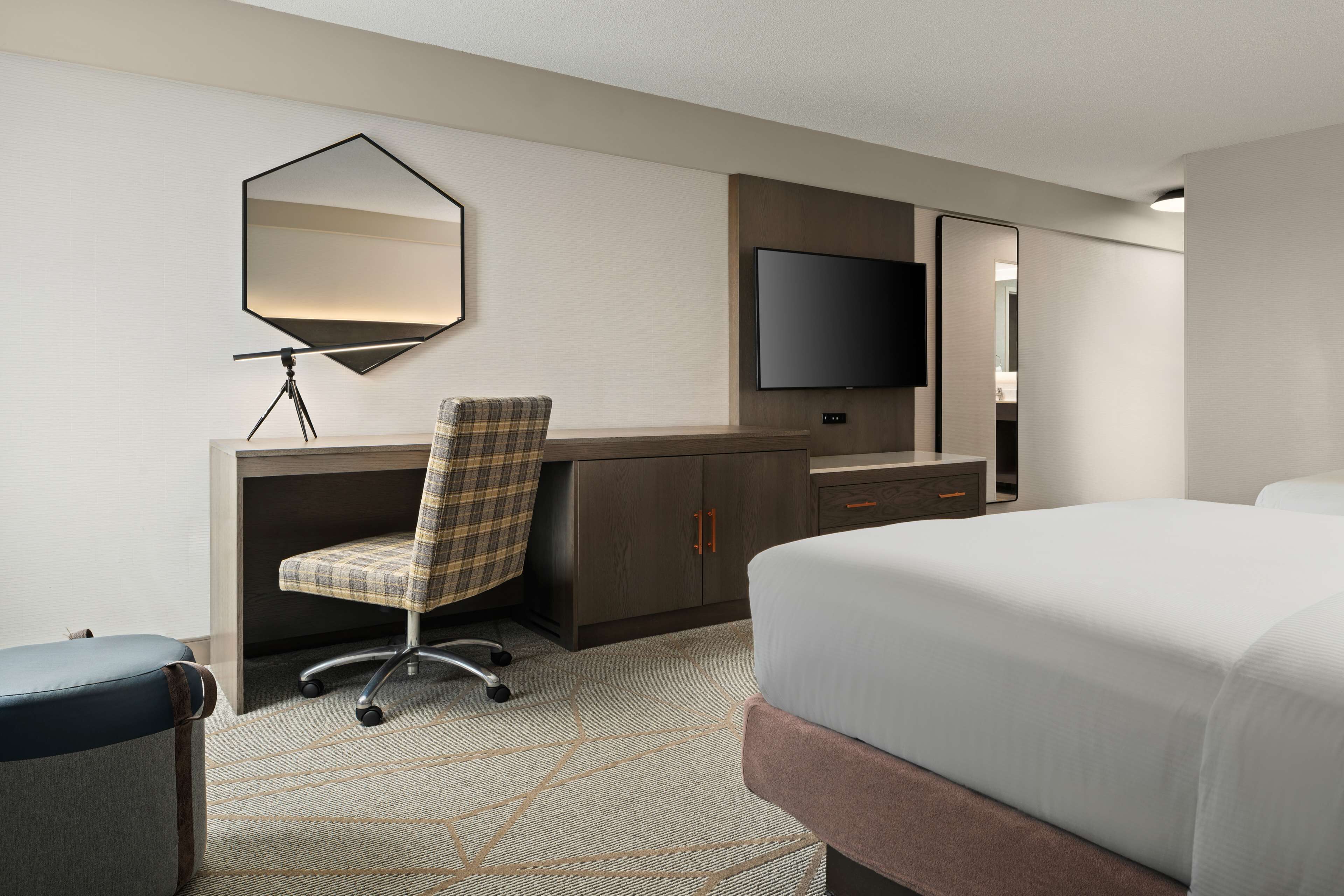 A hotel room with a bed, a desk with a chair, a flat-screen TV, a mirror, and a lamp.