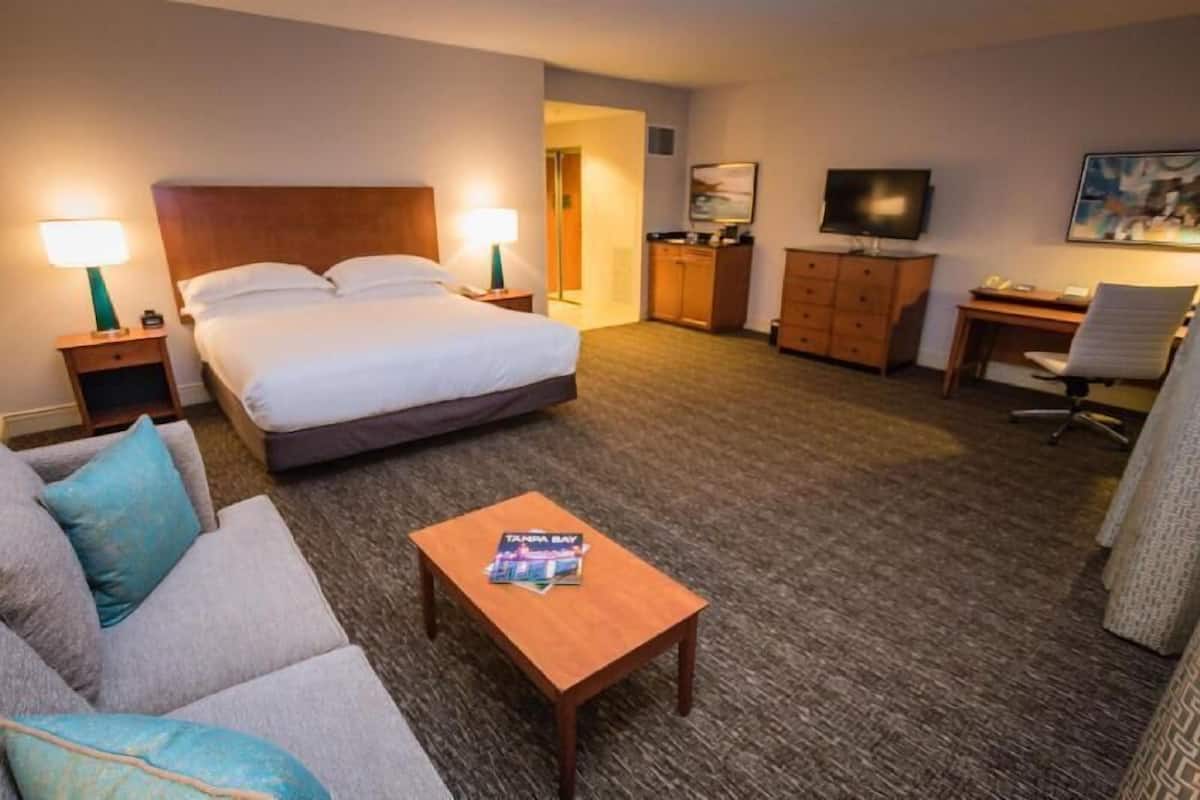 A hotel room with a bed, two bedside lamps, a TV, a desk, a chair, and a coffee table.